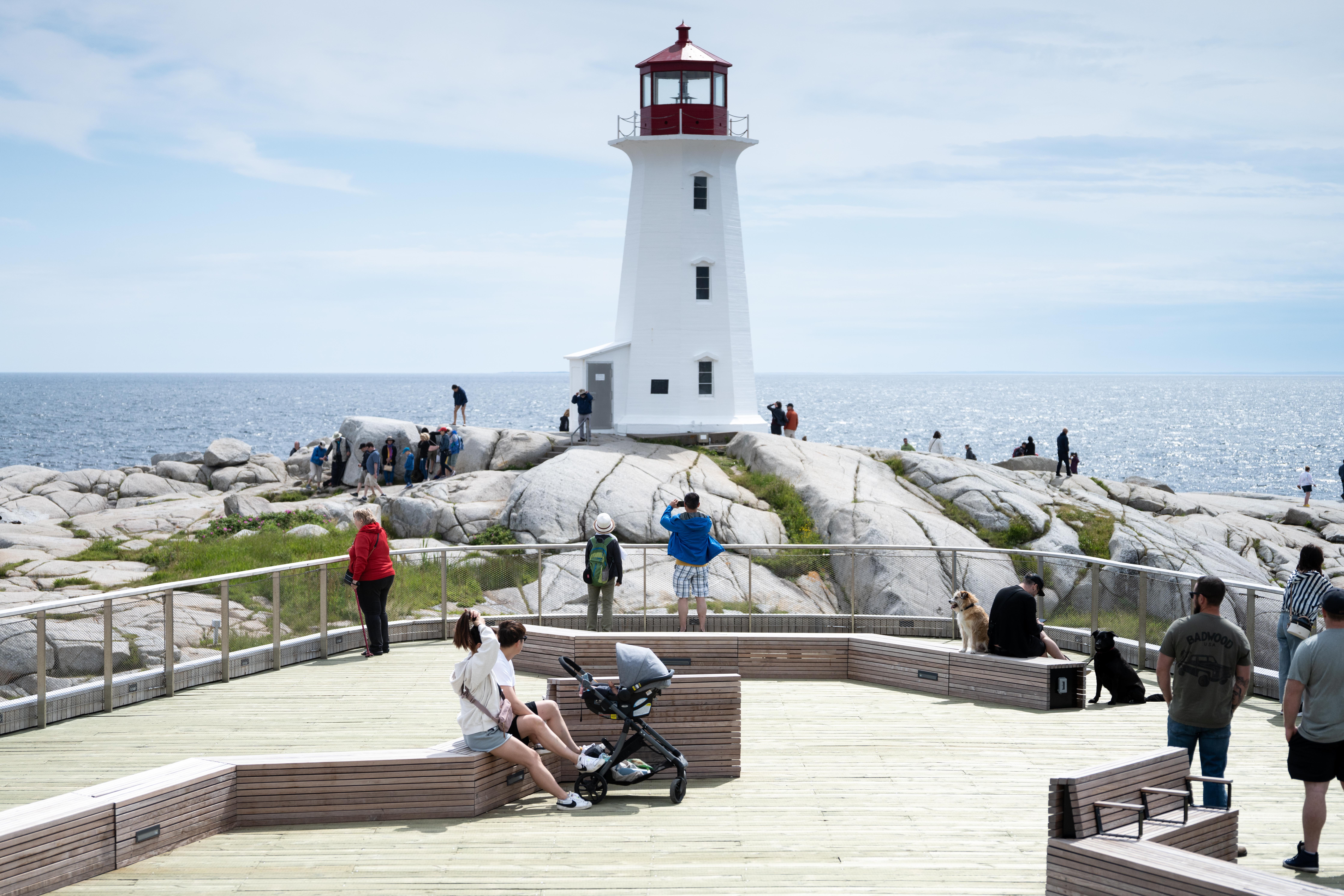 Front of the Peggy's Cove viewing deck. There are many people using the viewing deck, which is in close proximity to the lighthouse. The lighthouse is white and red and is built on large rocks overlooks the ocean