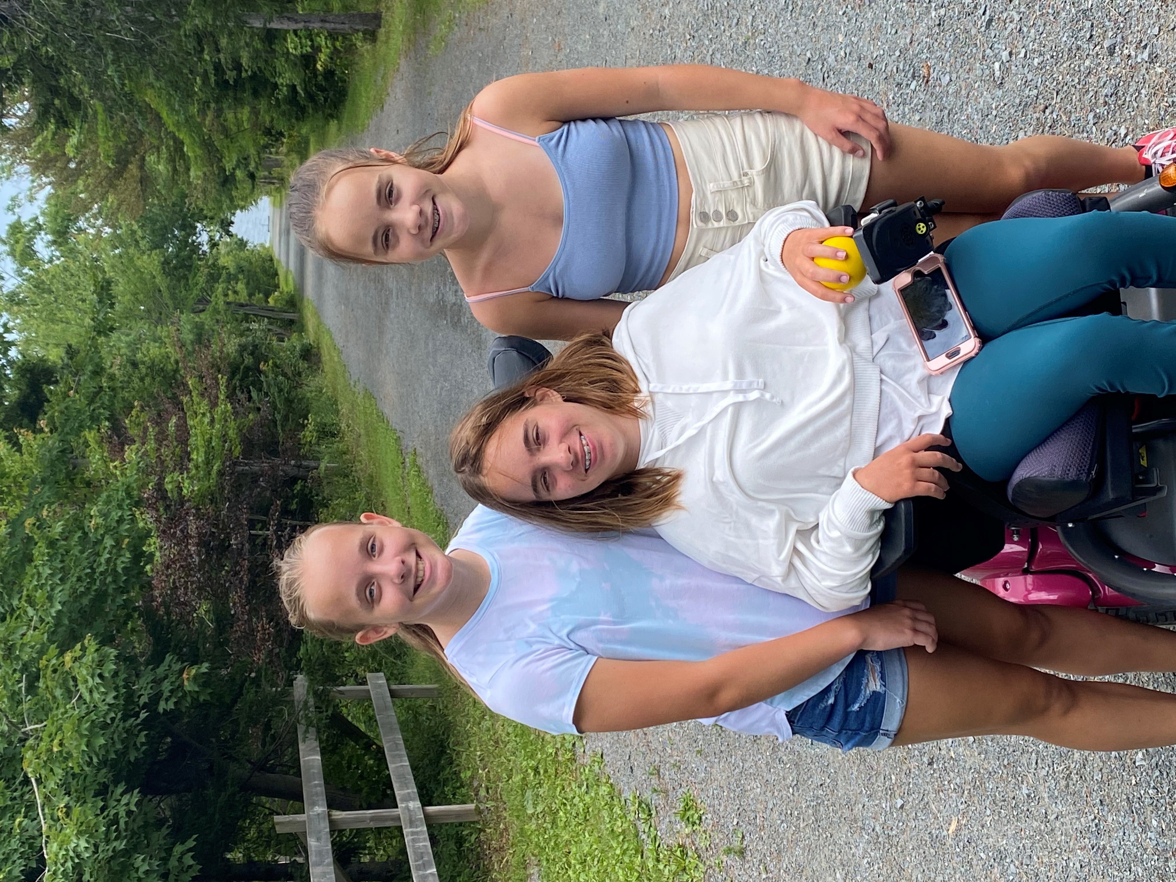 three girls outdoors on a cement path, one using a wheelchair, all smiling