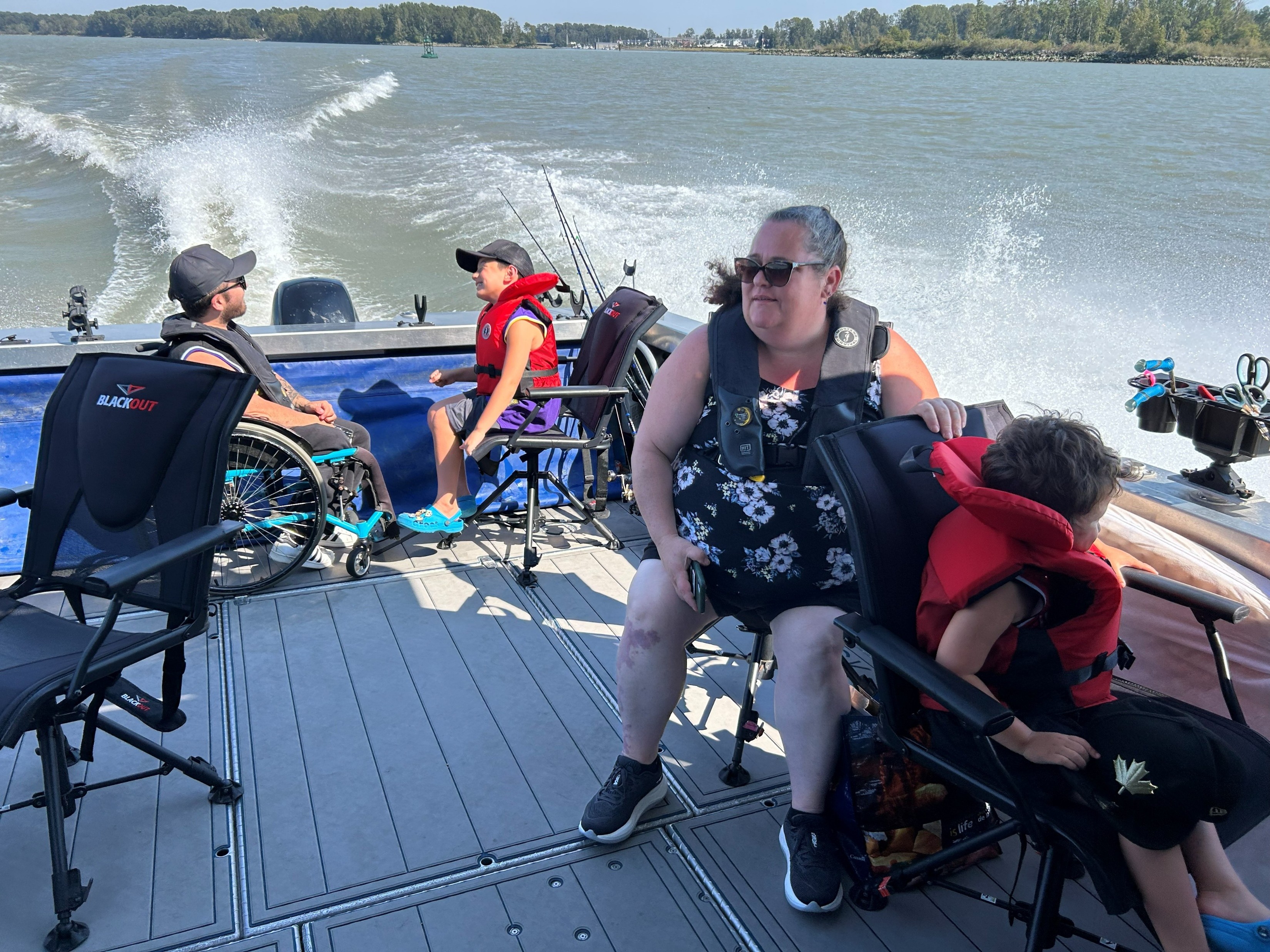 Group seated on an open boat travelling across the water, including two children wearing life jackets and one person using a wheelchair, with fishing rods secured behind them.