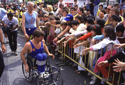 Rick Hansen on his wheelchair wheeling past youth supporters reaching over a metal fence to try and high-five him during the Man In Motion World Tour.