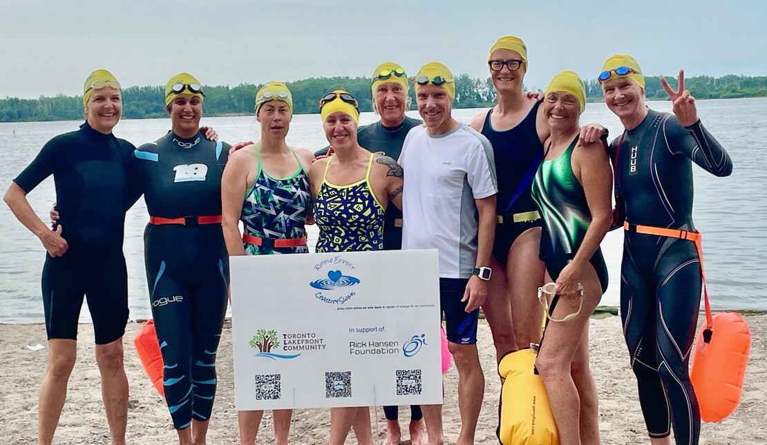 A group of swimmers wearing wetsuits and yellow swim caps stand together on a sandy beach near a lake. They are holding a large white sign featuring logos for “Toronto Event Community” and “Rick Hansen Foundation,”.