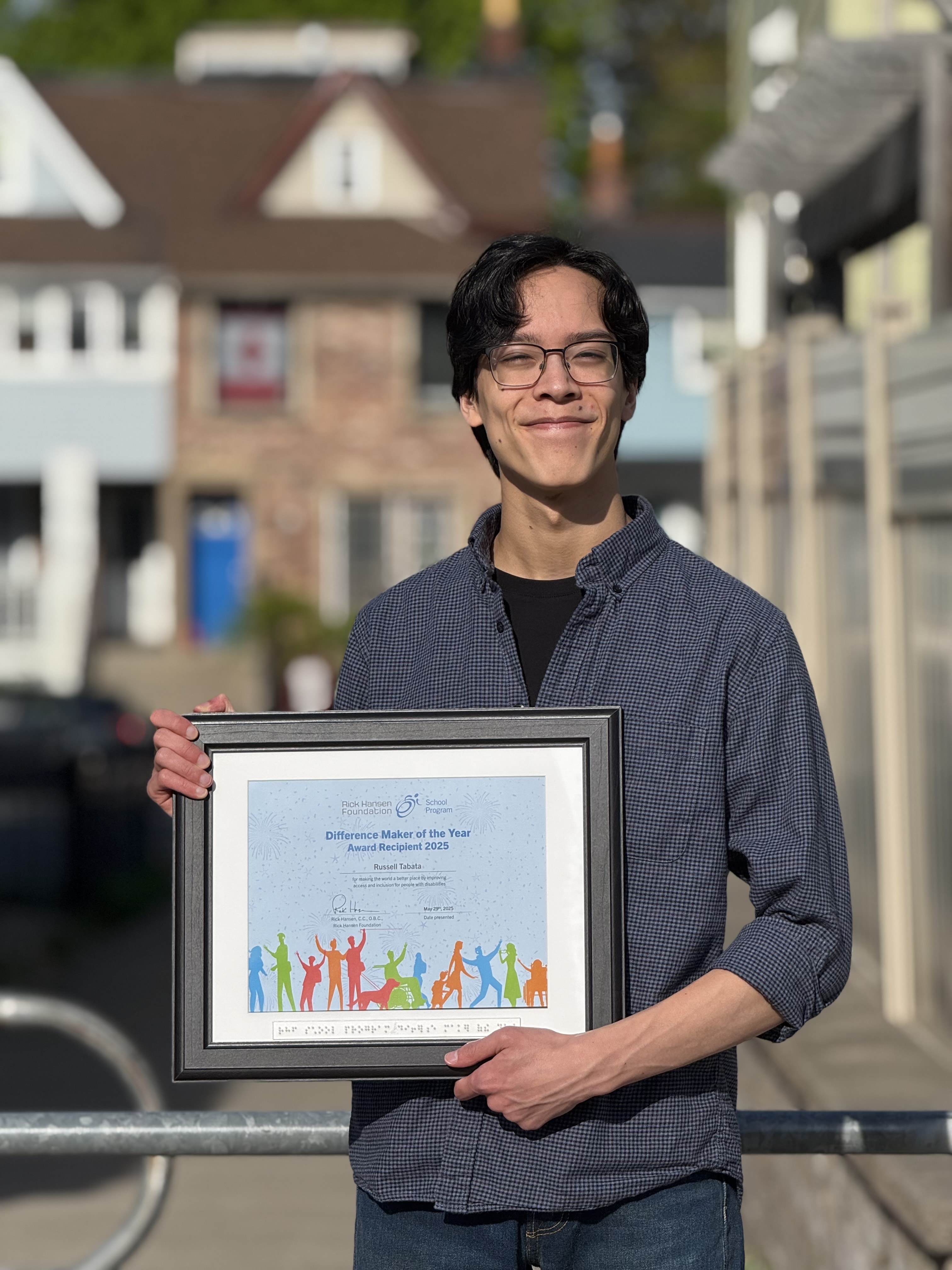 Smiling young man wearing glasses and holding a "Difference Maker of the Year Award Recipient 2025" certificate, with blurred houses in the background