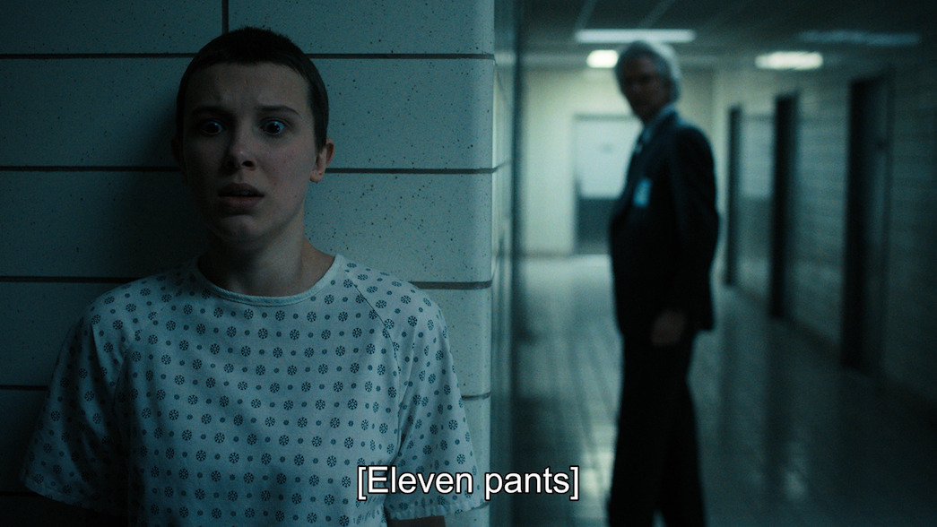 Eleven from stranger things, who has short dark hair, wearing a hospital gown and hiding behind a wall with a distressed expression. There is blurred image of someone wearing a black suit on the other side of of the wall. Closed captioning at the bottom reads "Eleven pants".