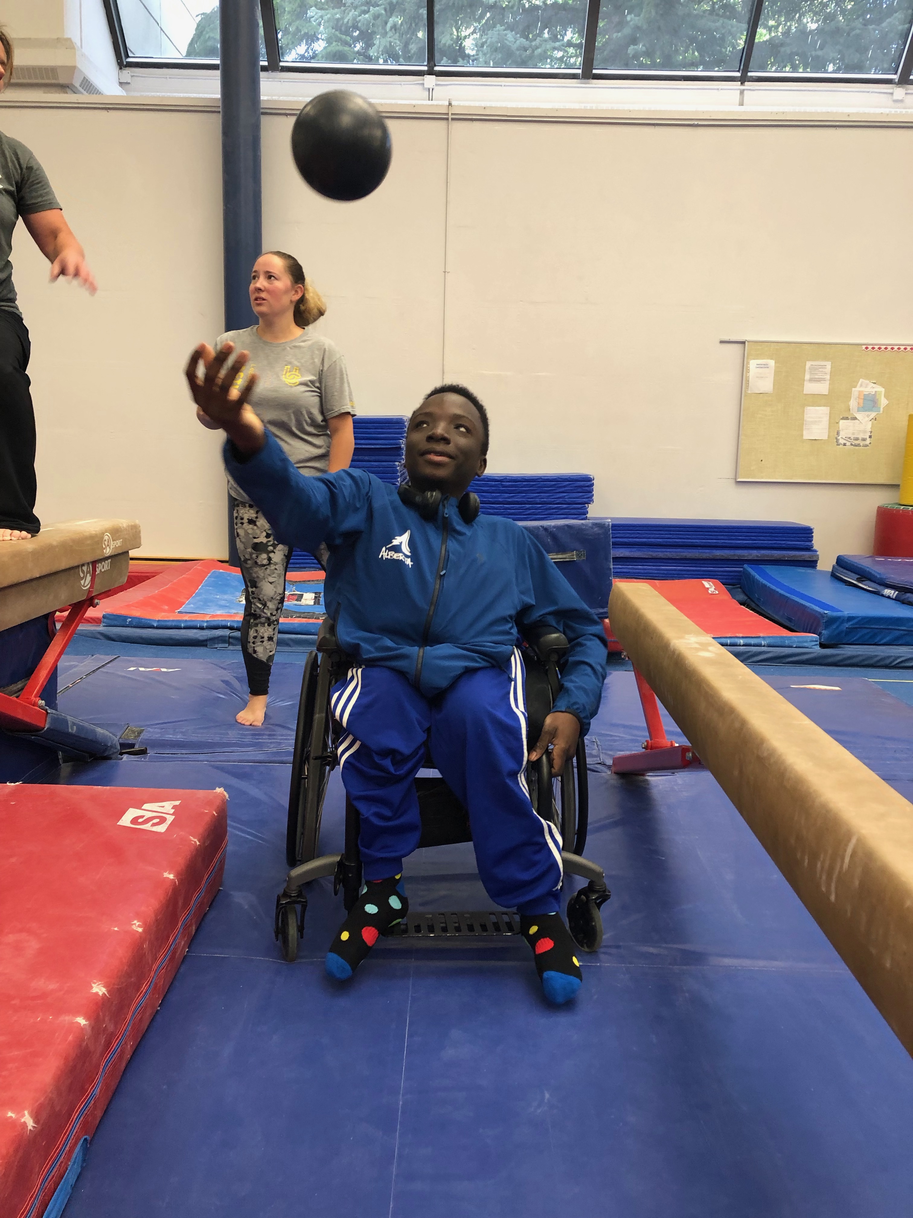 Young boy using a wheelchair and throwing a black medicine ball in the air. He is in a gymnasium.