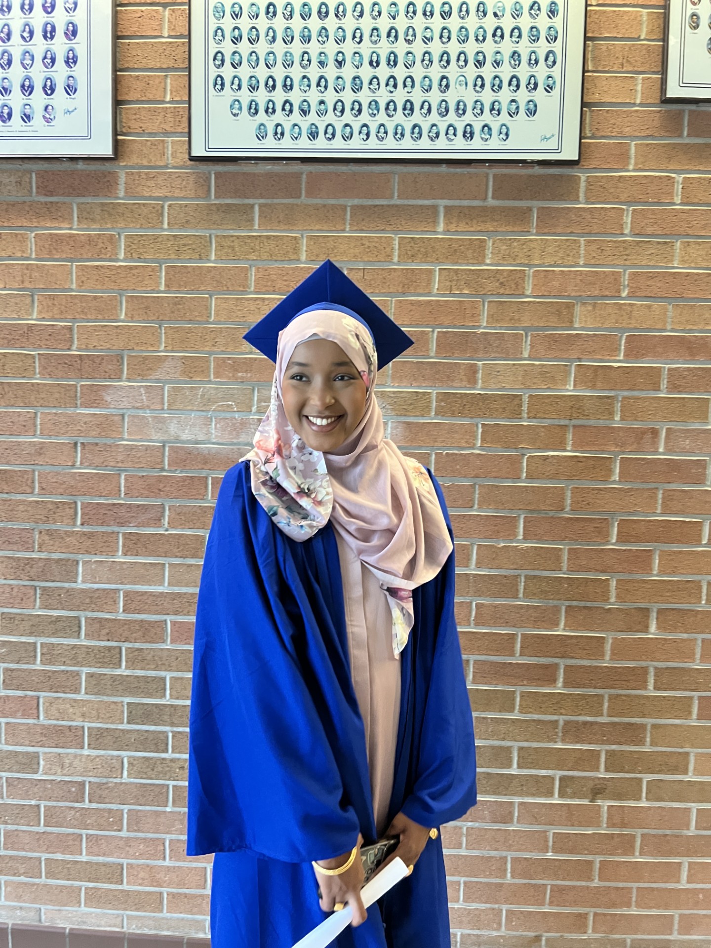 Young lady graduate wearing a blue cap and gown stands indoors holding a diploma while smiling at the camera