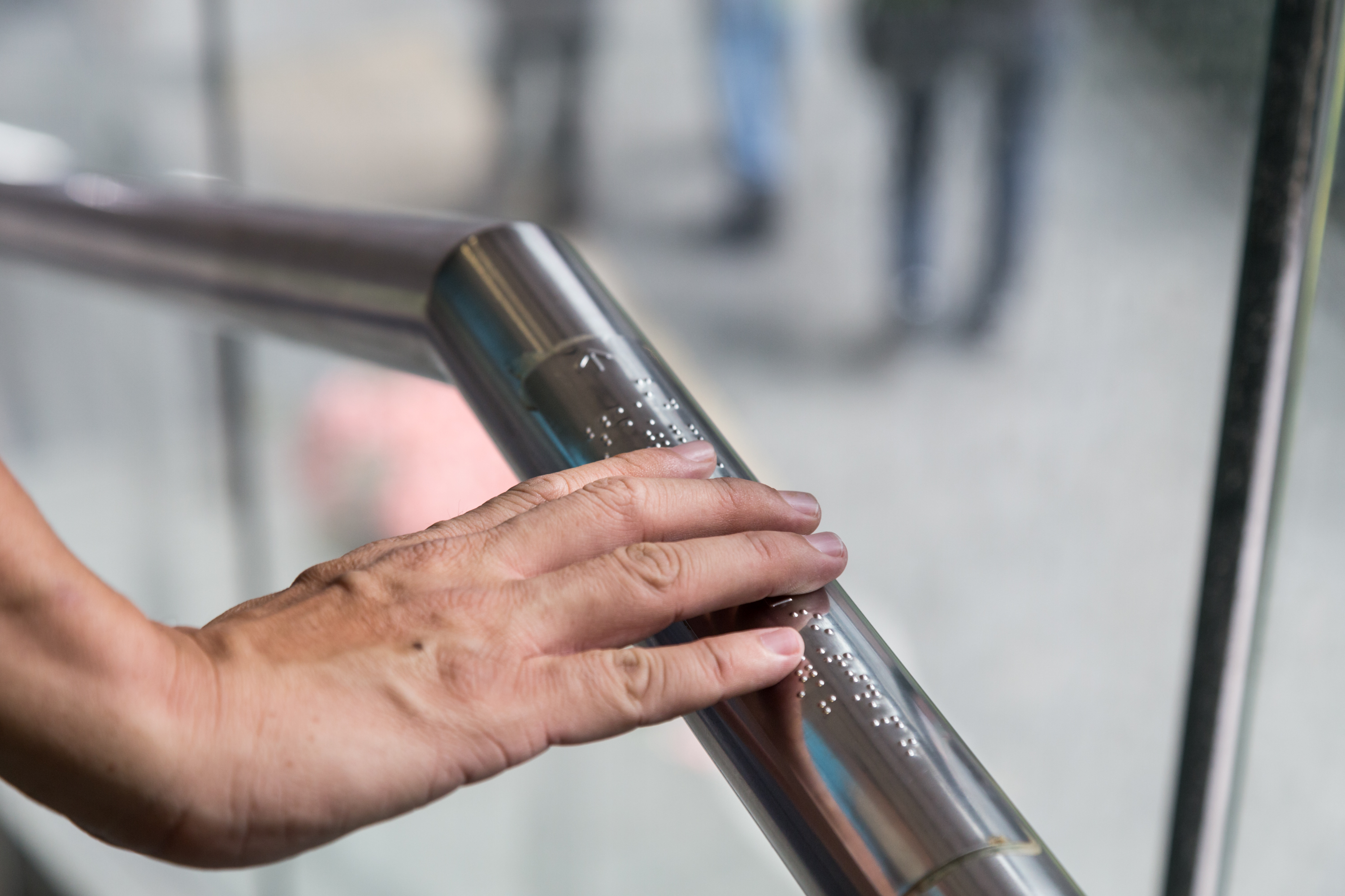 A hand reading Braille text on a metal railing inside a building.