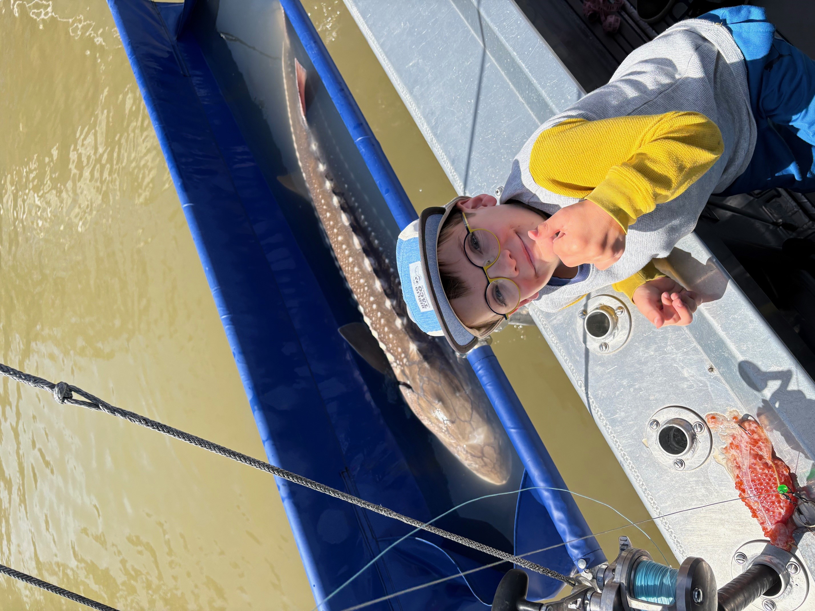 A boy on a boat next to a large fish in a blue container, with fishing gear visible.