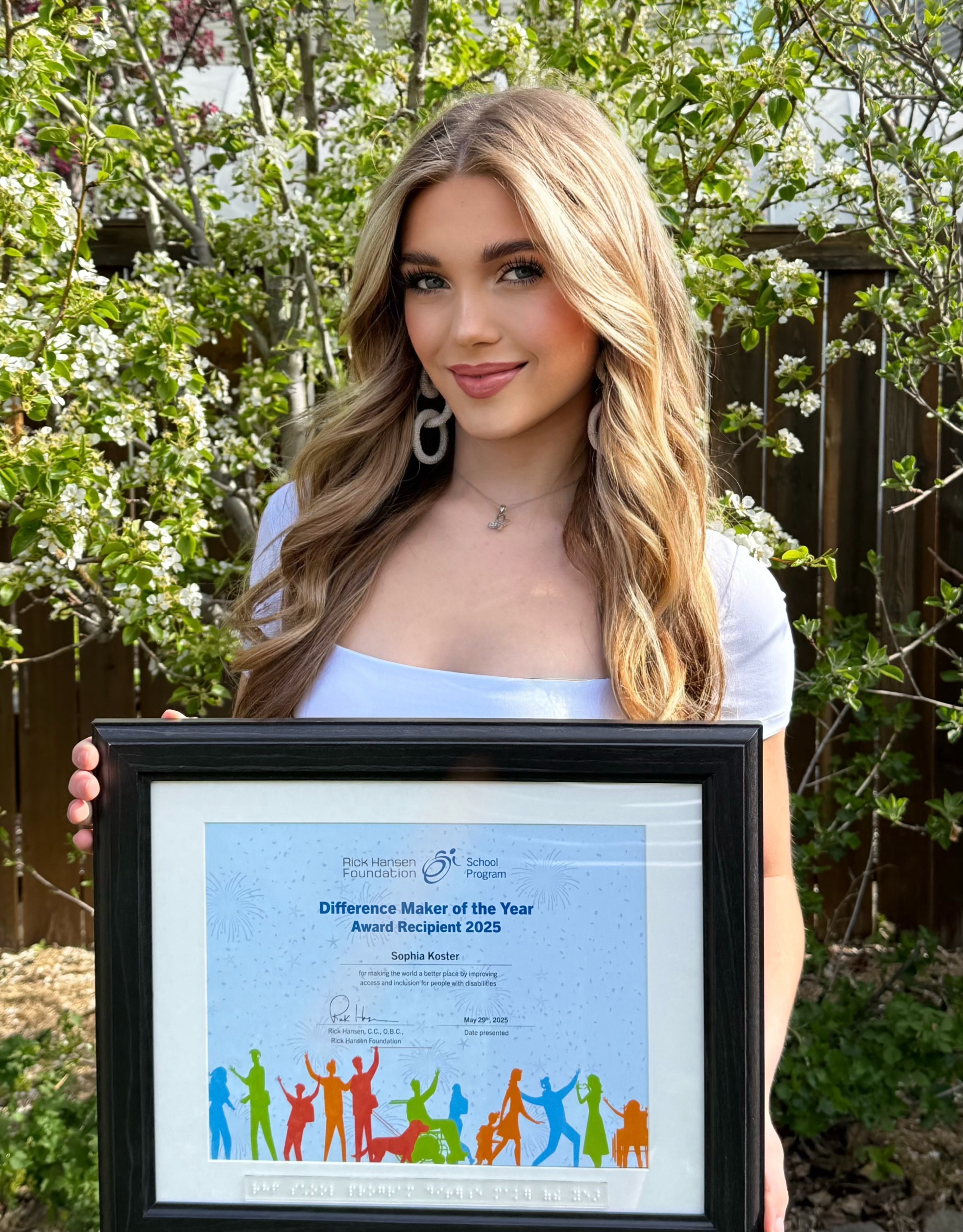 Young white woman smiling and holding a "Difference Maker of the Year Award Recipient 2025" certificate, with bushes in the background