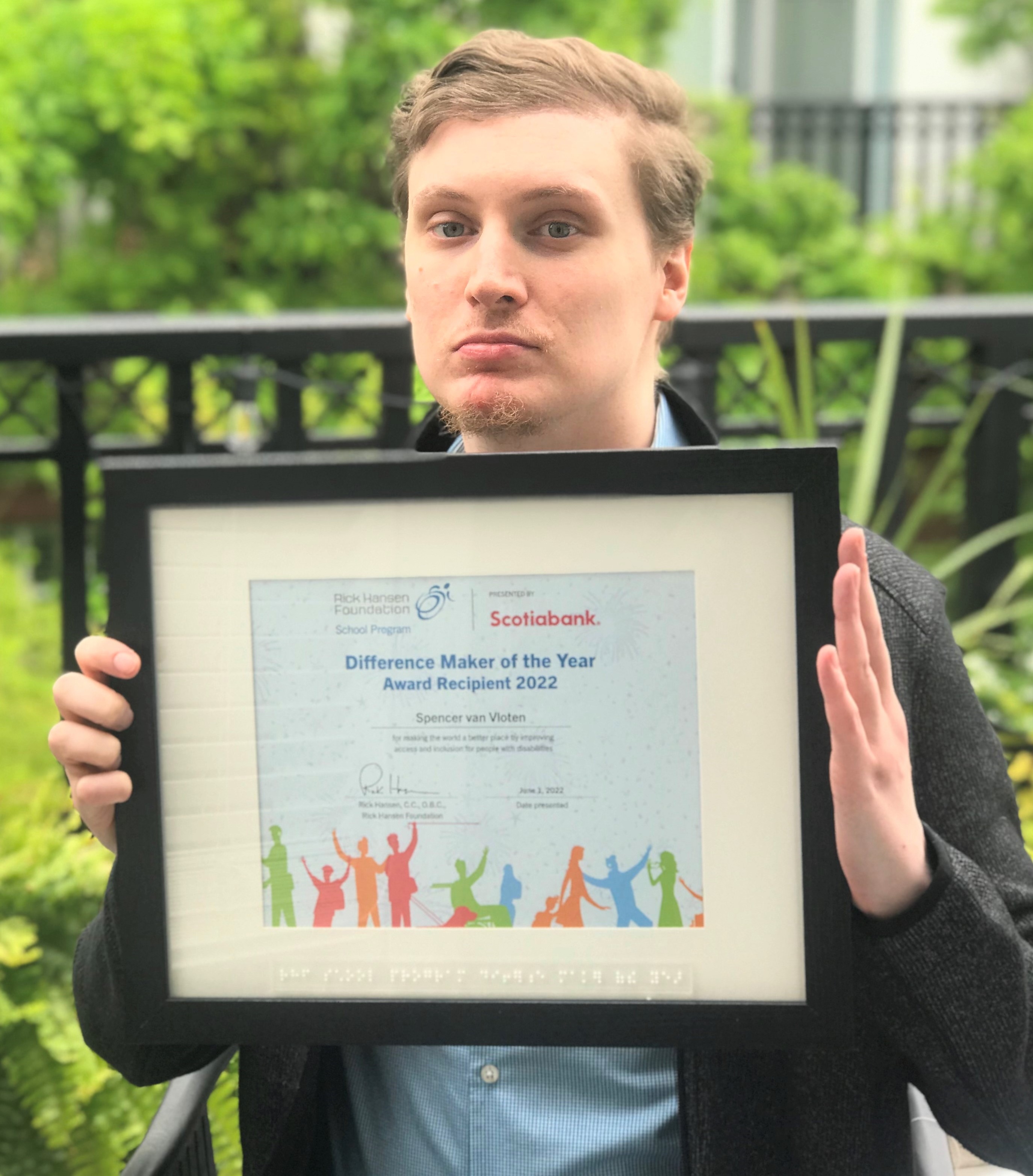 Young white adult man with light hair hold a framed difference maker award. He is sitting in front of trees with green leaves.