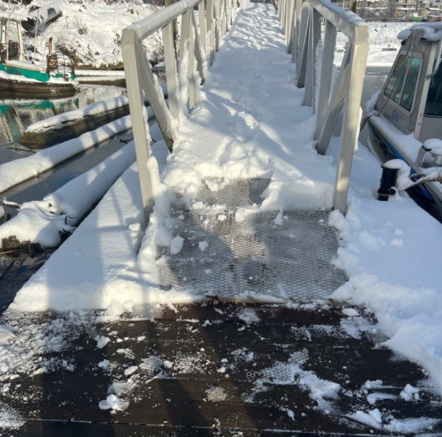Snow-covered dock or ramp leading over water, with metal railings on both sides and footprints visible in the snow. Boats are partially visible along the sides.