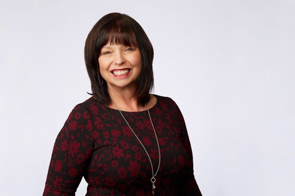 Woman with brown hair, wearing red floral pattern shirt smiles for the camera.