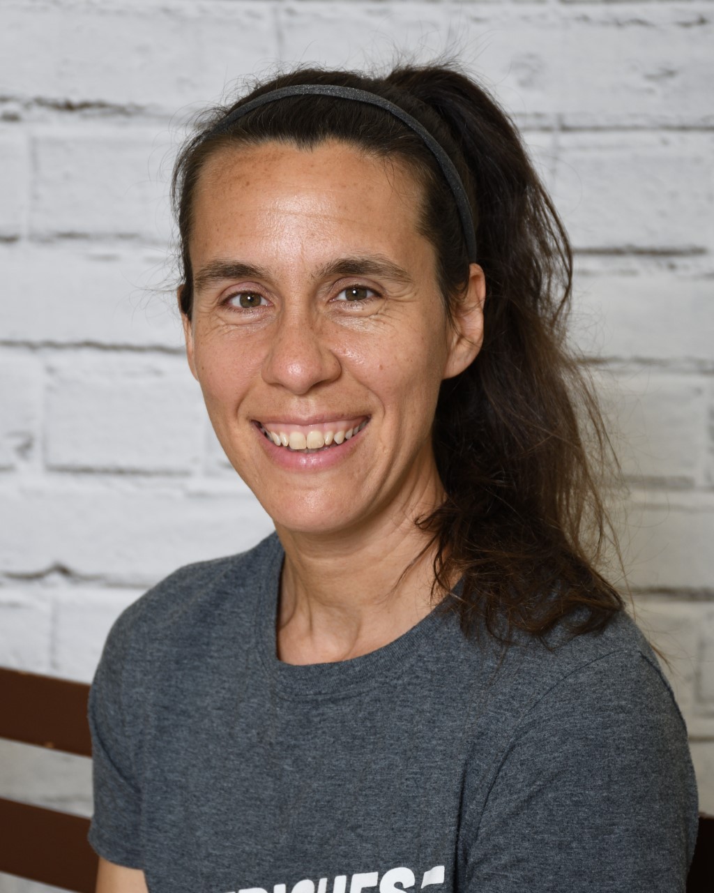 Woman wearing a grey t-shirt smiling. She has dark brown hair in a ponytail. 