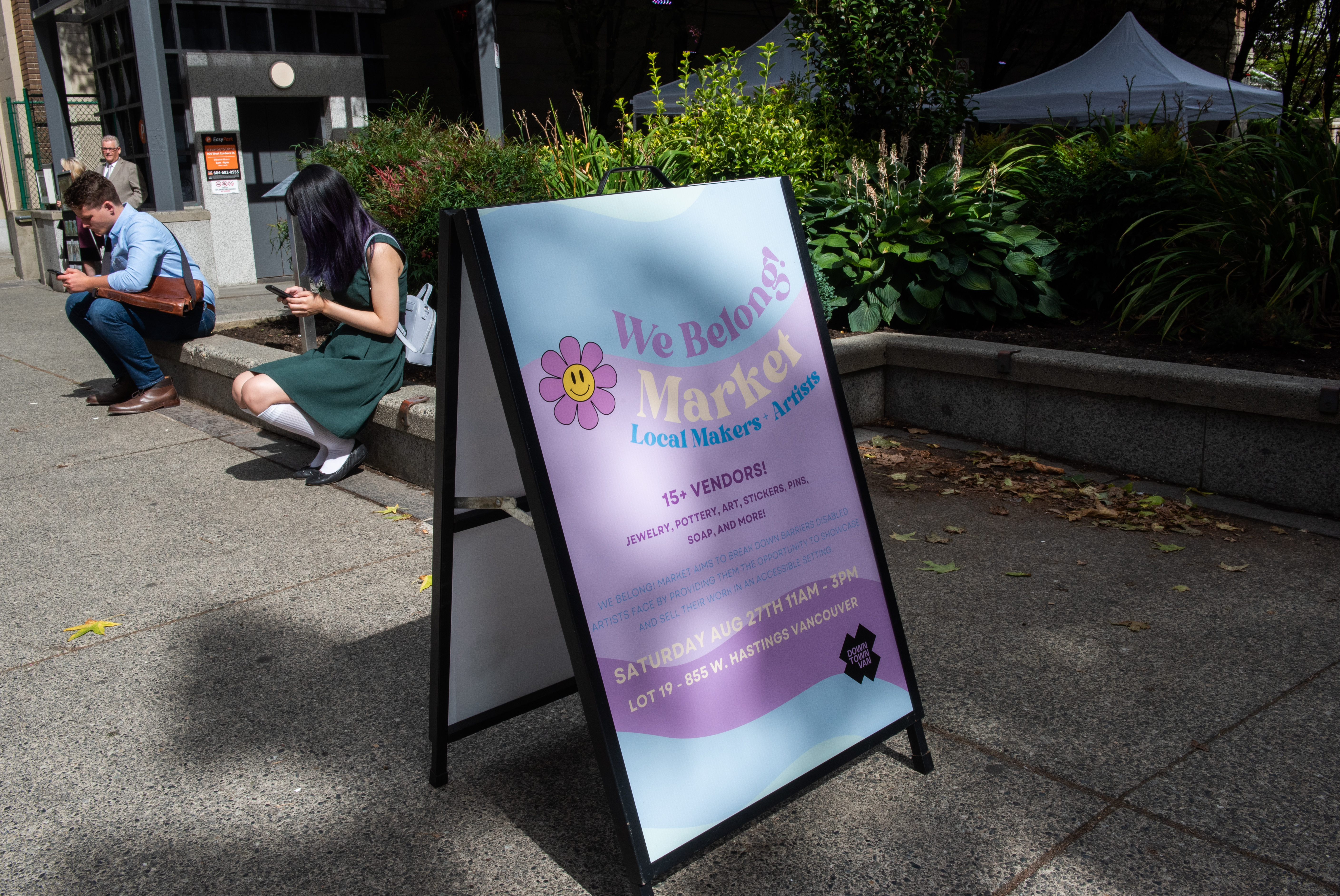 Blue and purple sign that reads "We Belong! Market Local Makers + Artists. 15+ Vendors! Jewelry, Pottery, Art, Stickers, Pins, Soap and More! We Belong! Market aims to break down barriers disabled artists face by providing them the opportunity to showcase and sell their own in an accessible setting. Saturday Aug. 27th 11AM - 3 PM Lot 19-  855 W. Hastings Vancouver