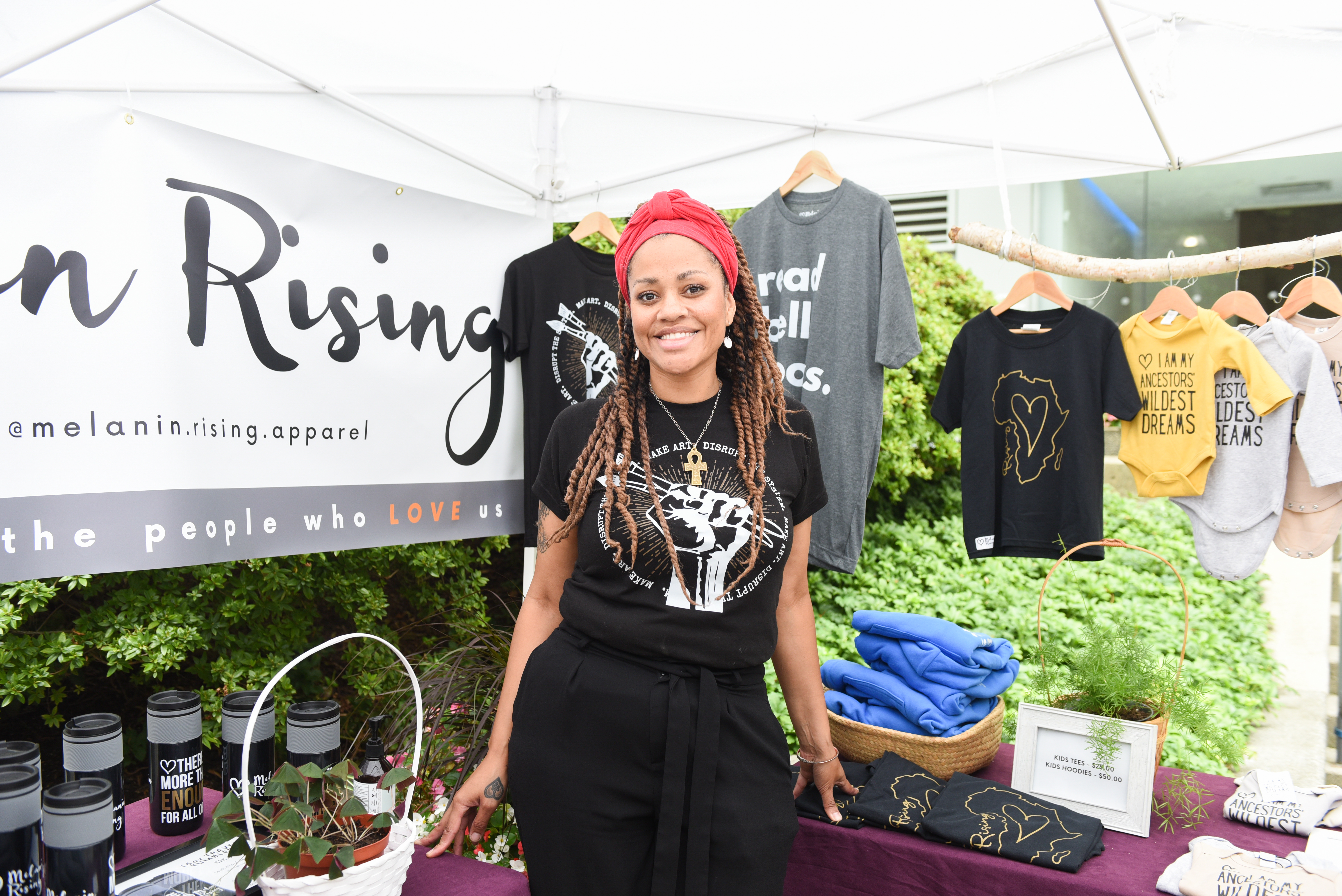 Naomi Grace of Melanin Rising who has long braids and is wearing a black t-shirt with a fist on it that is holding two paint brushes. Naomi is standing in her the Melanin Rising booth.
