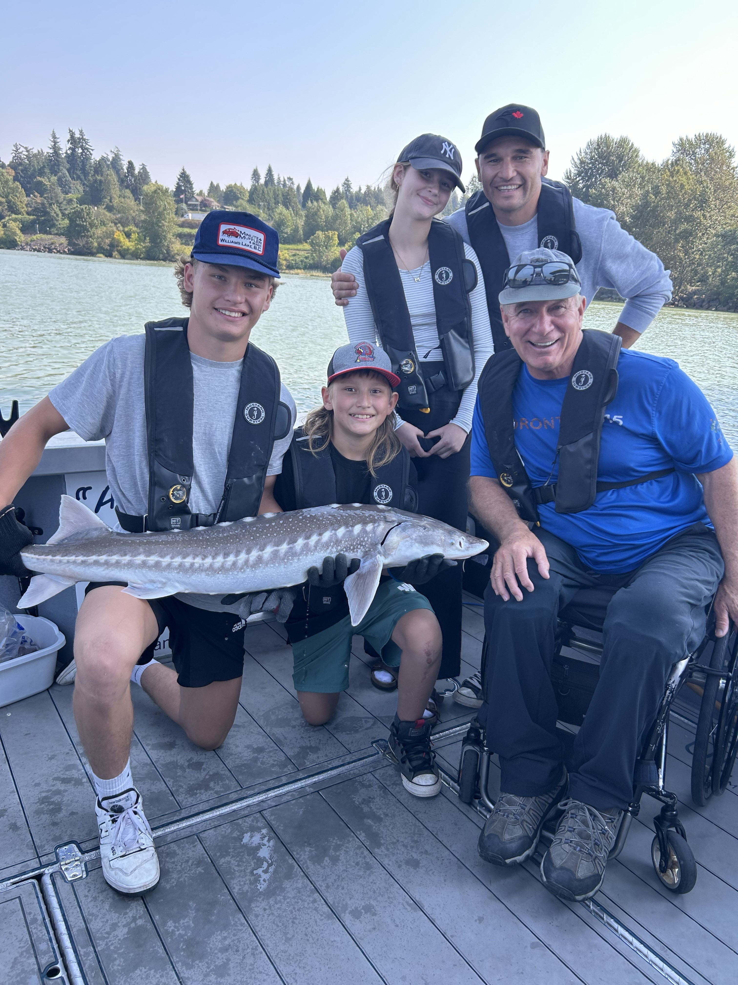 Group gathered on a fishing boat displaying a large fish. One person (Rick) is using a wheelchair, and all are wearing life jackets on the water.