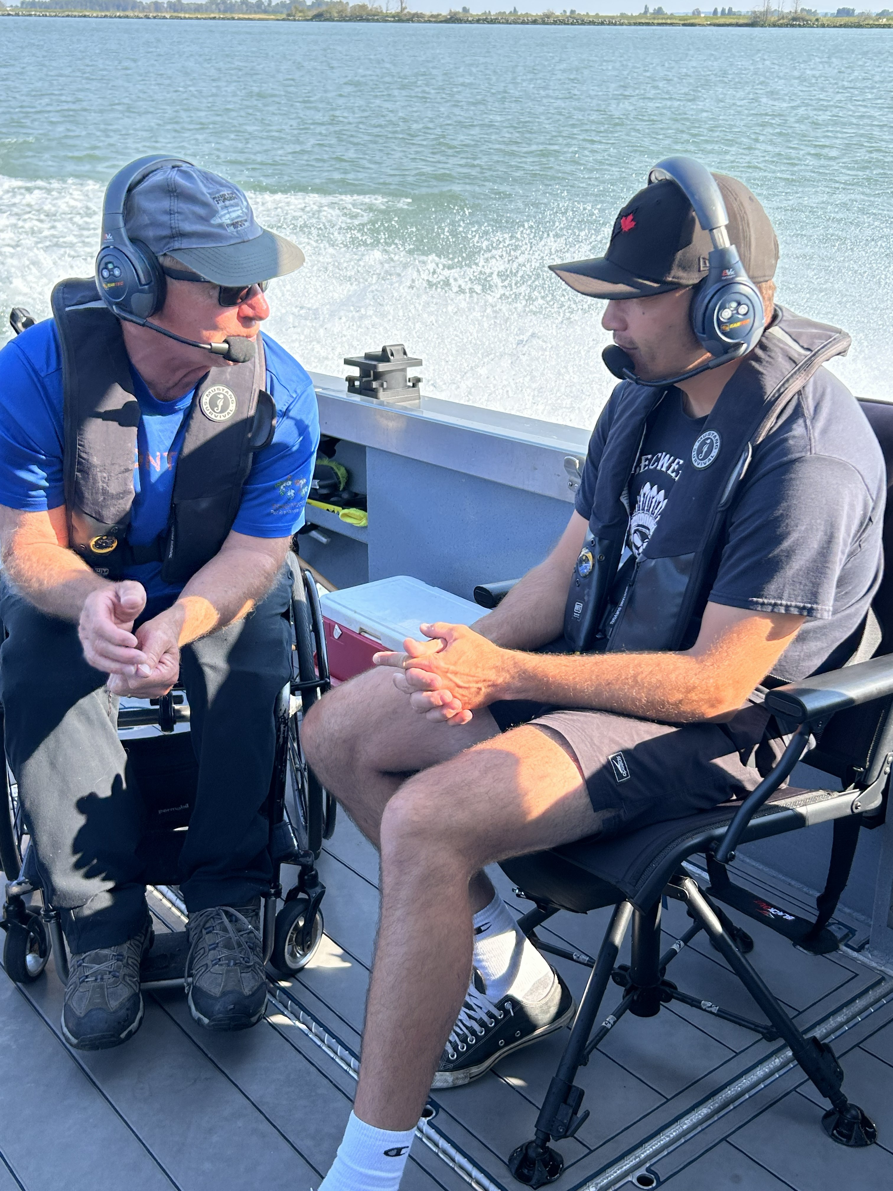 Two people seated on an accessible fishing boat wearing headsets and life jackets, talking while traveling across the water.