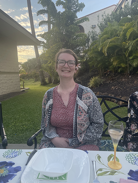 Alana is seated at an outdoor dining table surrounded by blue skies, palm trees, and a building. She is wearing a floral decoration on her hair, glasses, and smiling in a summer floral outfit.