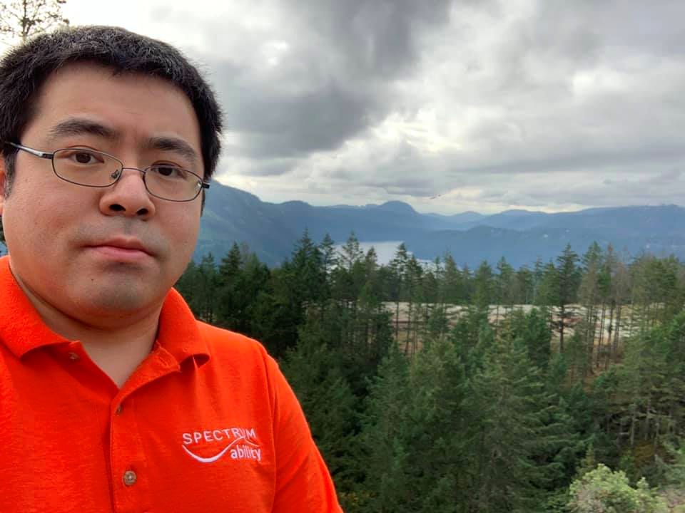 Man with black hair and glasses in an orange t-shirt that says Spectrum Ability in white in the corner. The background is trees and mountains.
