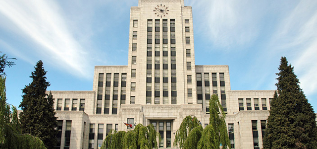 Vancouver City Hall, a large beige building with a clock at the top. It is surrounded by trees.