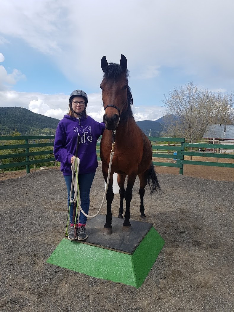 Elise stands beside her brown horse on a pedestal, her horse has two front hooves on the pedestal.  She is wearing a helmet, purple hoodie, blue jeans and running shoes. She is holding her horse by the leash. They are in a stable with mountains in the background.