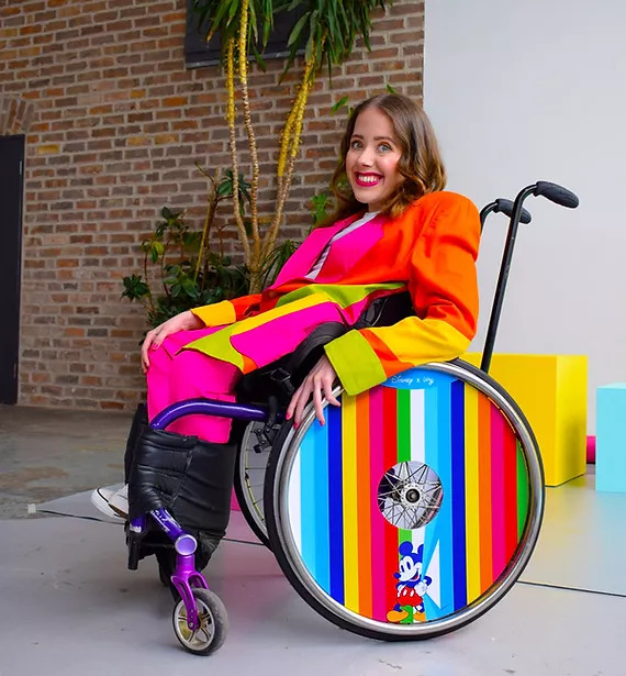A wheelchair model is dressed in bright orange blazer and pink pants, while her chair wheels are multicoloured in blue, cyan, yellow, red, and green stripes. A blue Mickey Mouse is a the bottom of her wheel. Behind her is an office with trees and white and bricked walls.