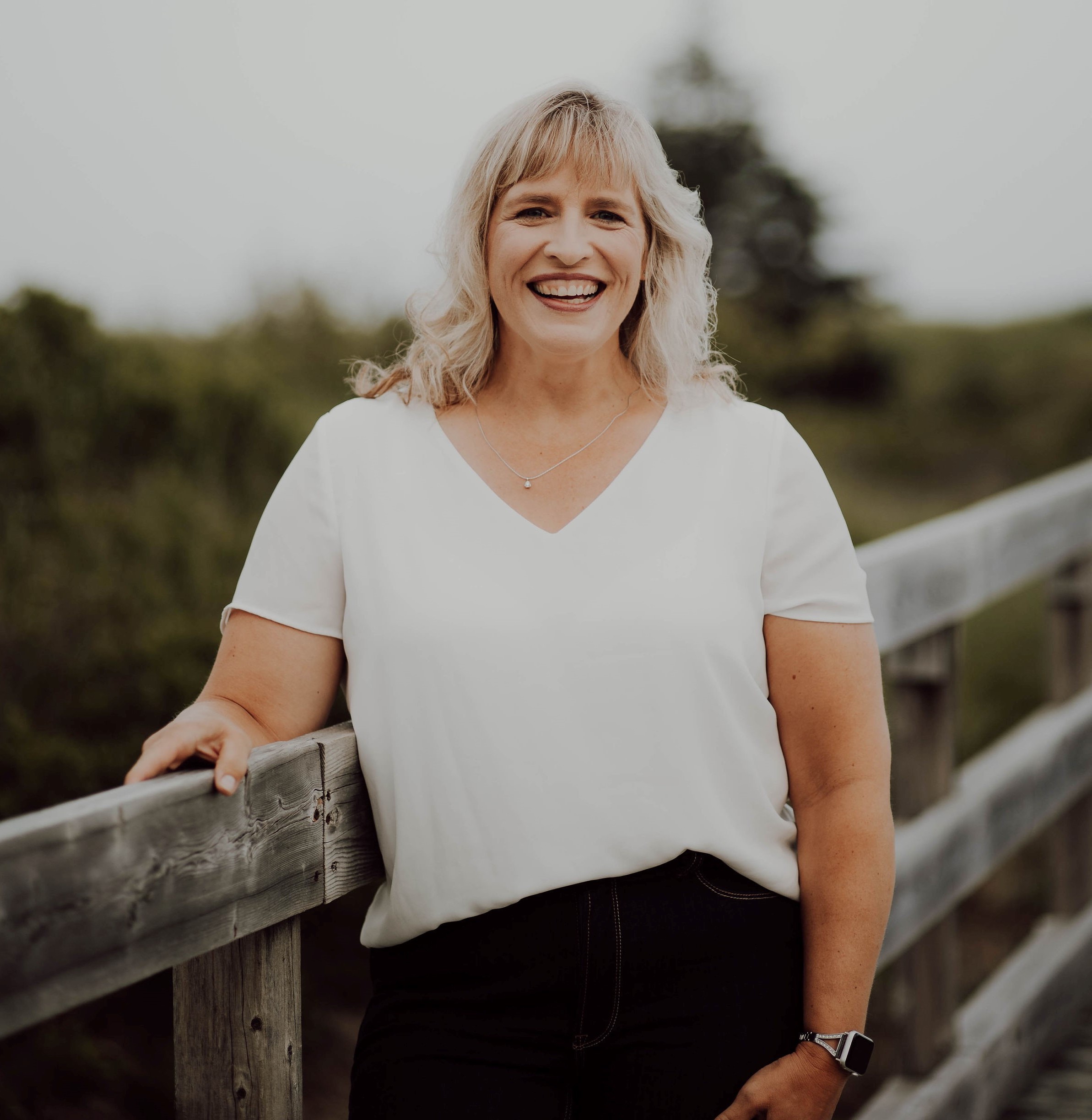 Woman with blonde hair standing in front of a wooden fence outside. She is wearing a white t-shirt and black pants.