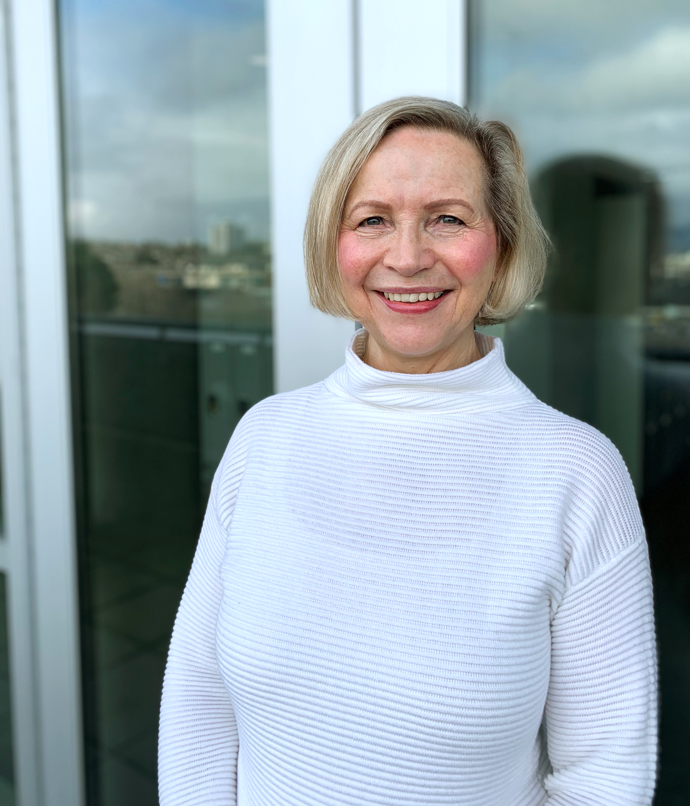 Pat Short wearing a white turtleneck sweater and smiling at the camera. Pat has chin-length light coloured hair.