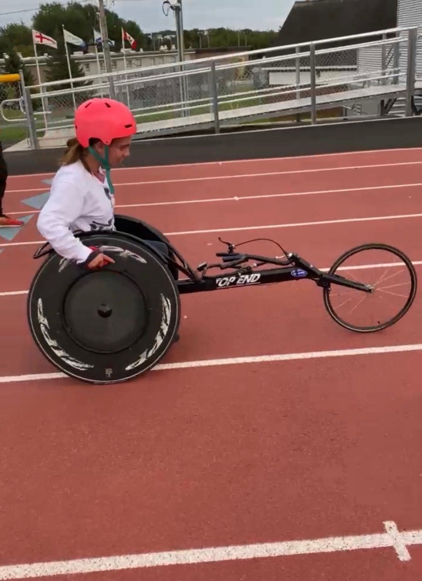 Payton Given is pictured on her racing wheelchair, with a pink helmet. She is on a track and field wheeling across.
