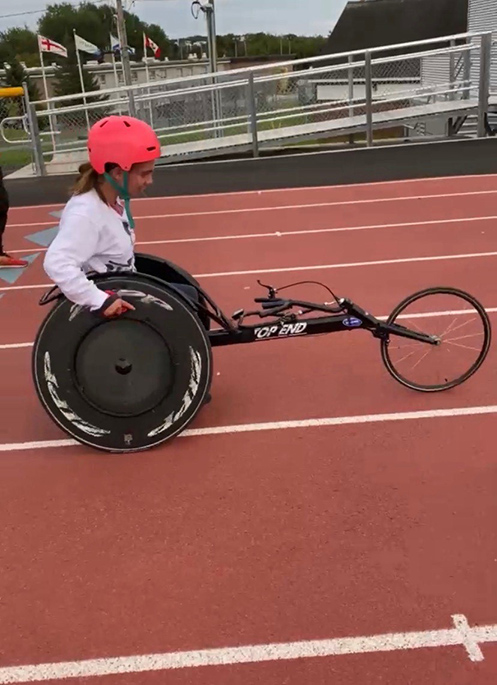 Payton is racing in a wheelchair on a track and field. She is wearing a pink helmet.