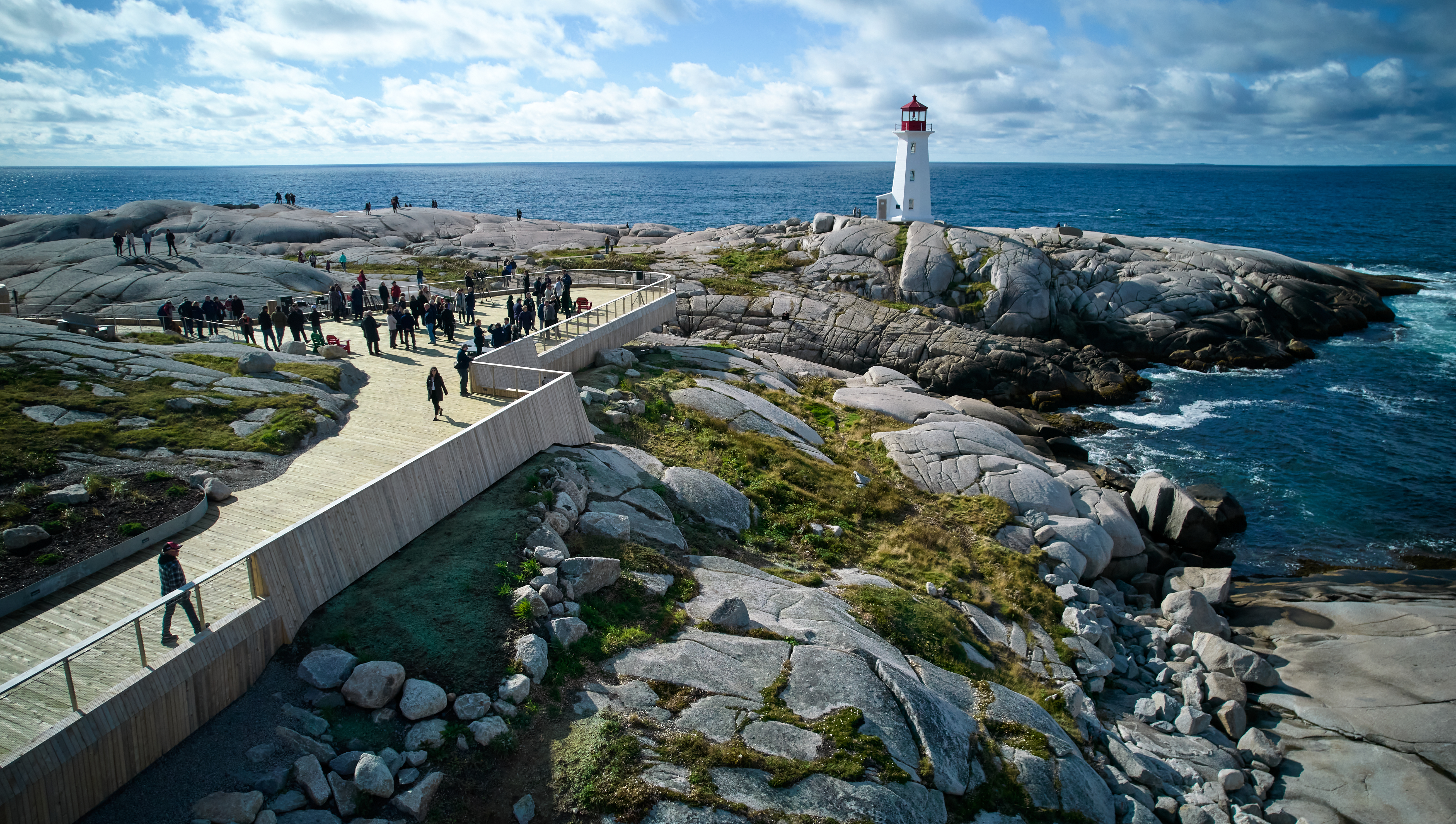 Aerial view of Peggy's Cove lighthouse. There is a large, wooden accessible viewing deck with a crowd of people towards the front. The viewing deck is built on top of large rocks. The lighthouse is red and white and overlooks the ocean.