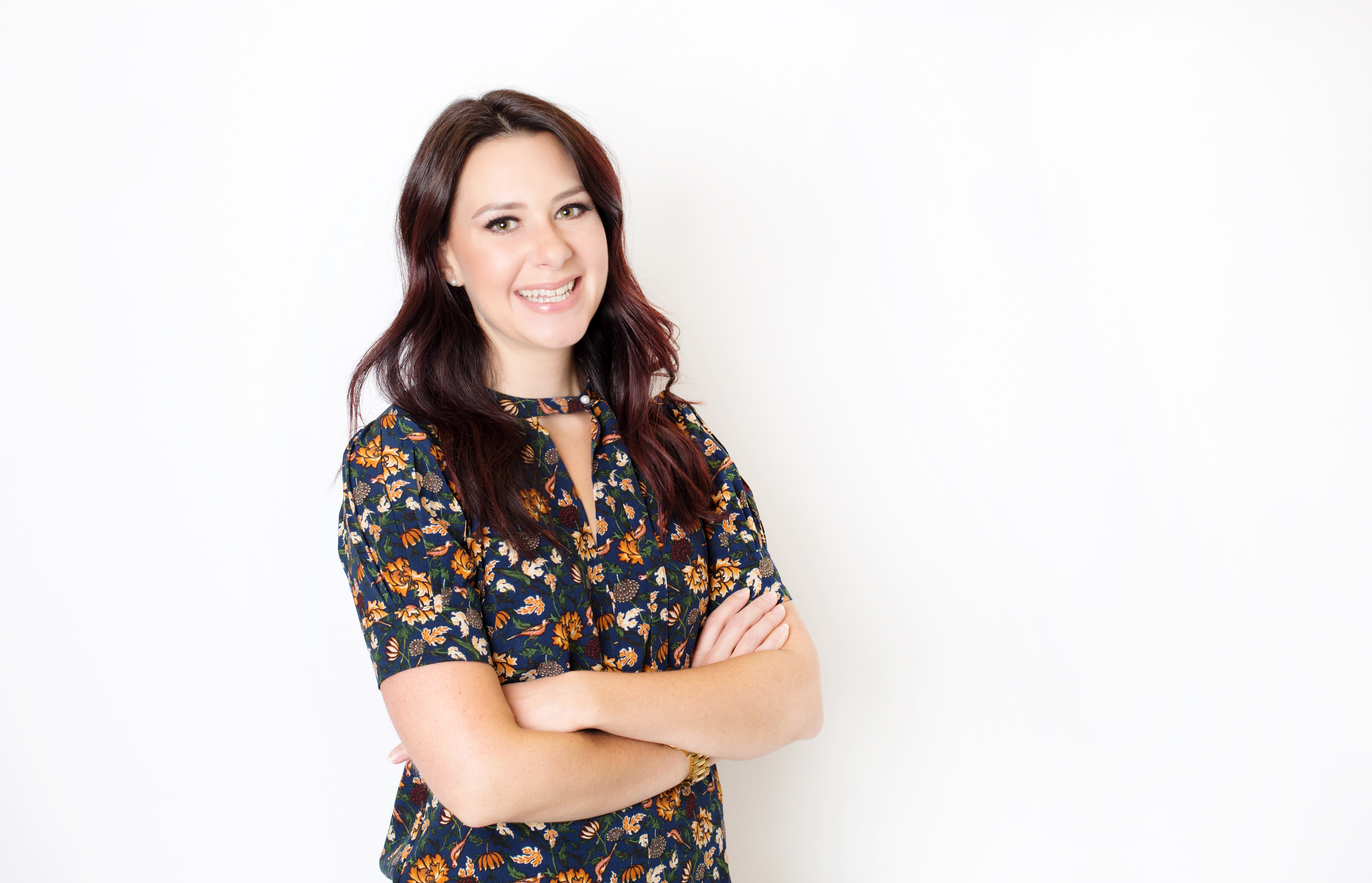 Samantha Ryan who is in front of a white background. Samantha has long dark hair and is wearing a dress with flowers on it.