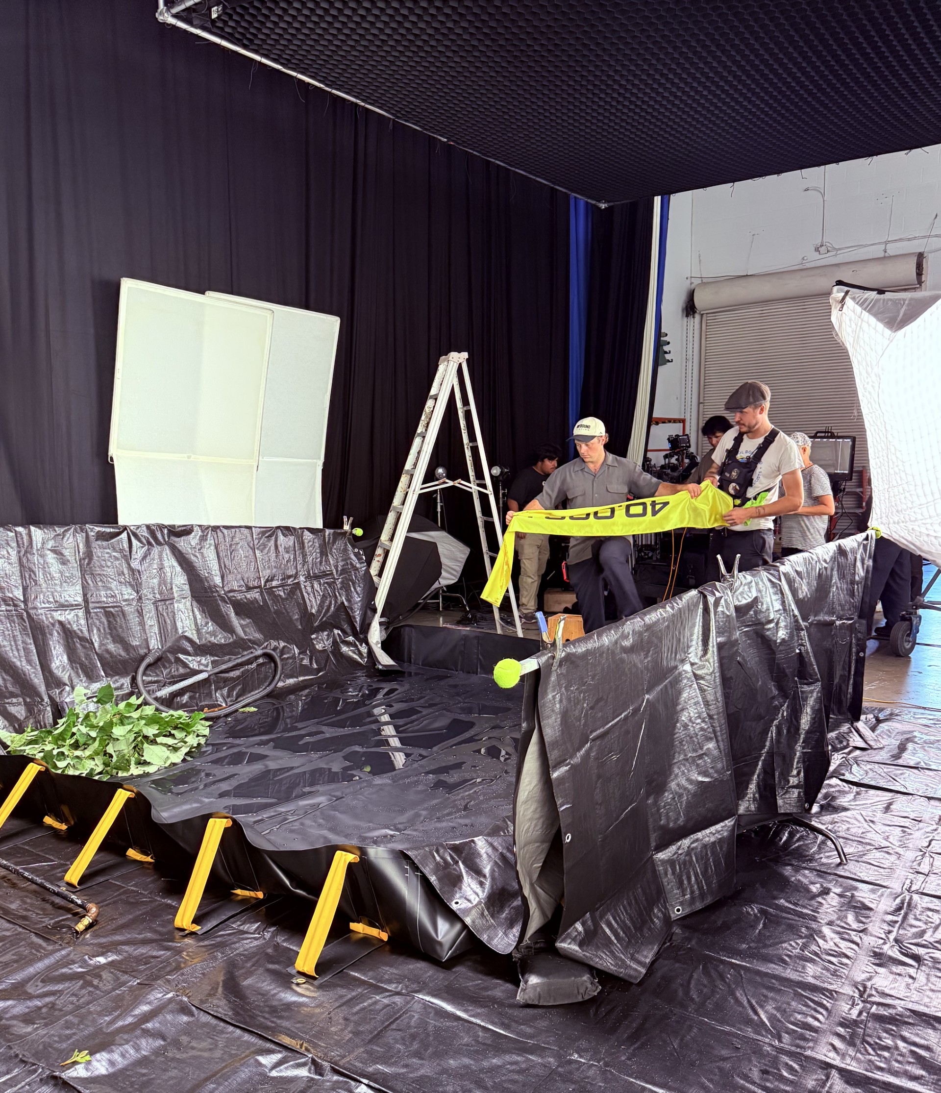 Behind-the-scenes studio set with crew members preparing a staged area covered in black material, surrounded by lighting equipment, cameras, and a ladder.