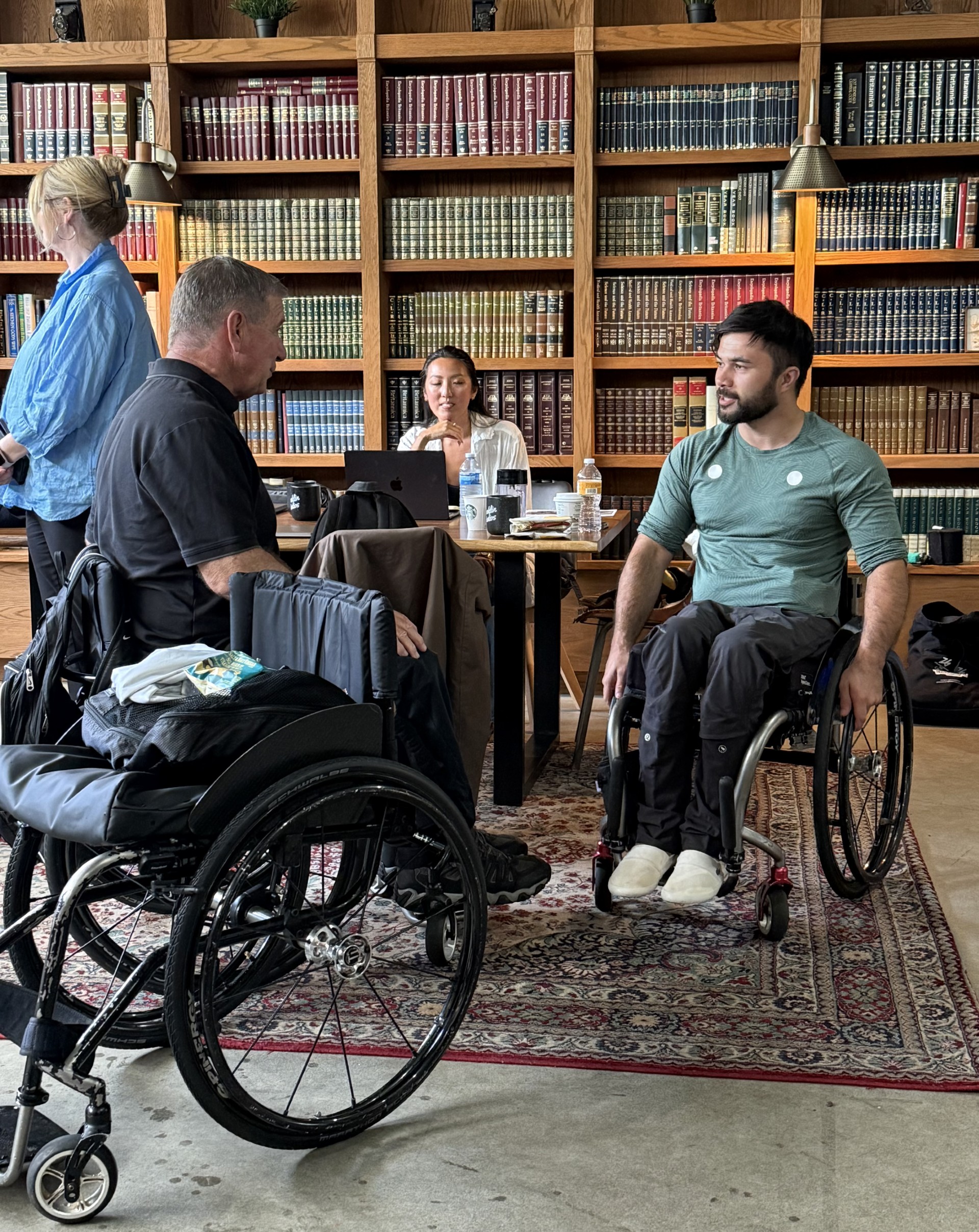 Two people in a wheelchair talking in a room with bookshelves in the background, while two other people are nearby