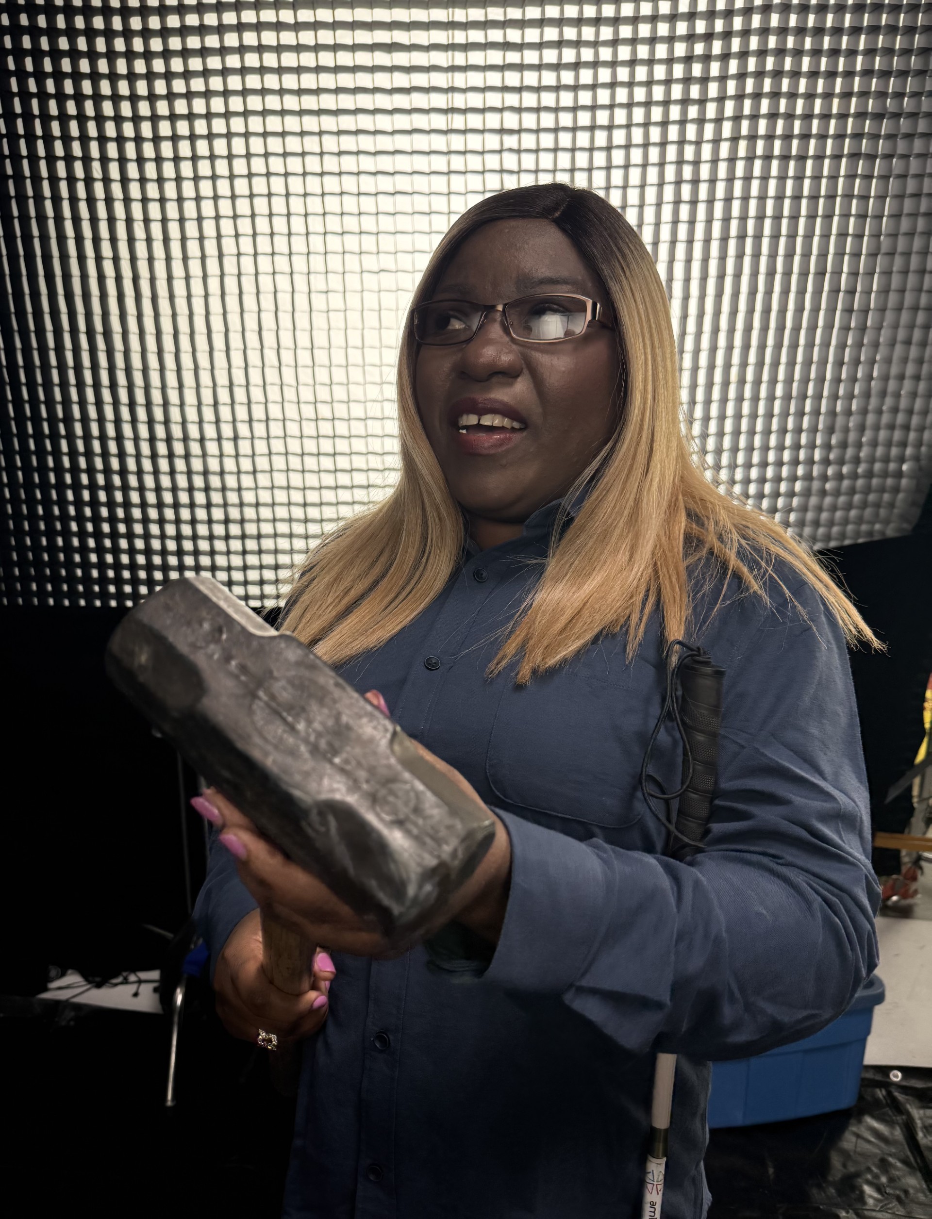 Woman with long blonde hair and glasses holding a large sledgehammer, with a white cane visible beside her, in a studio setting.