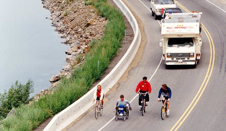 Rick Hansen using wheelchair and team on bicycles going through a road. 