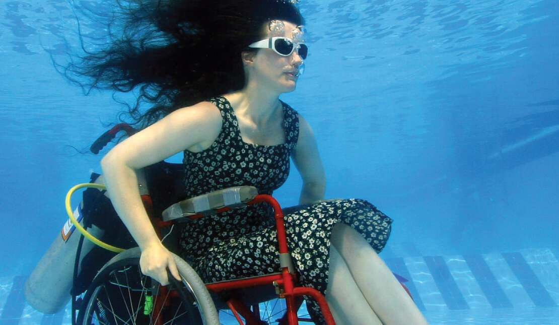 Woman in wheelchair underwater at bottom of pool with goggles 