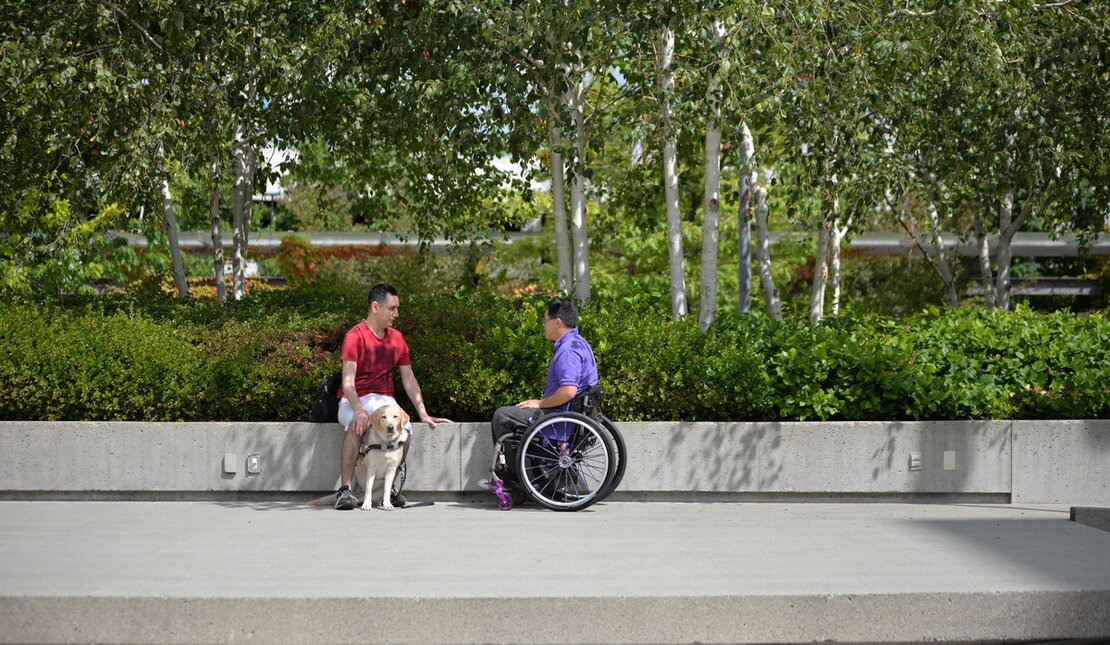 Man with seeing eye dog sits next to man using a wheelchair in a public park on a sunny day