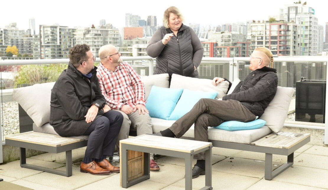 Front view of a group of 3 Caucasian men and 1 Caucasian women who are Deaf and Hard of Hearing people on lounge furniture in a rooftop patio. The woman is standing behind the furniture, centred, and signing to the men sitting down.  The background overlooks downtown Vancouver.