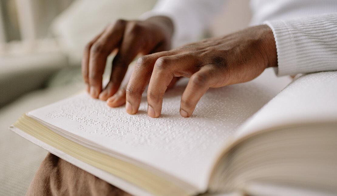 A braille book open on a person's lap, being read with their fingers, in a room full of natural light 