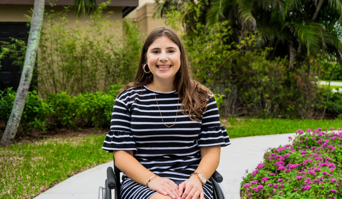 Girl with long brown hair sitting in a wheelchair outdoors, smiling and wearing a blue dress