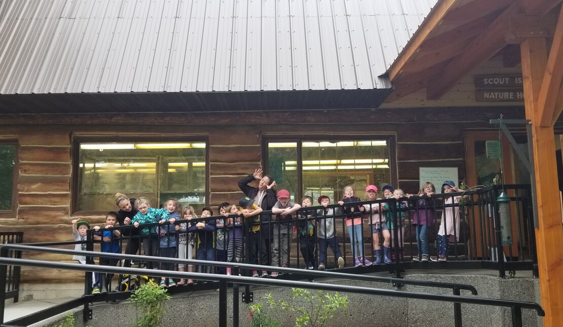 A group of children make funny faces with their teachers by the ramp railings outside Scout Island Nature Centre, a wooden structure with angled roof and and wide windows. 