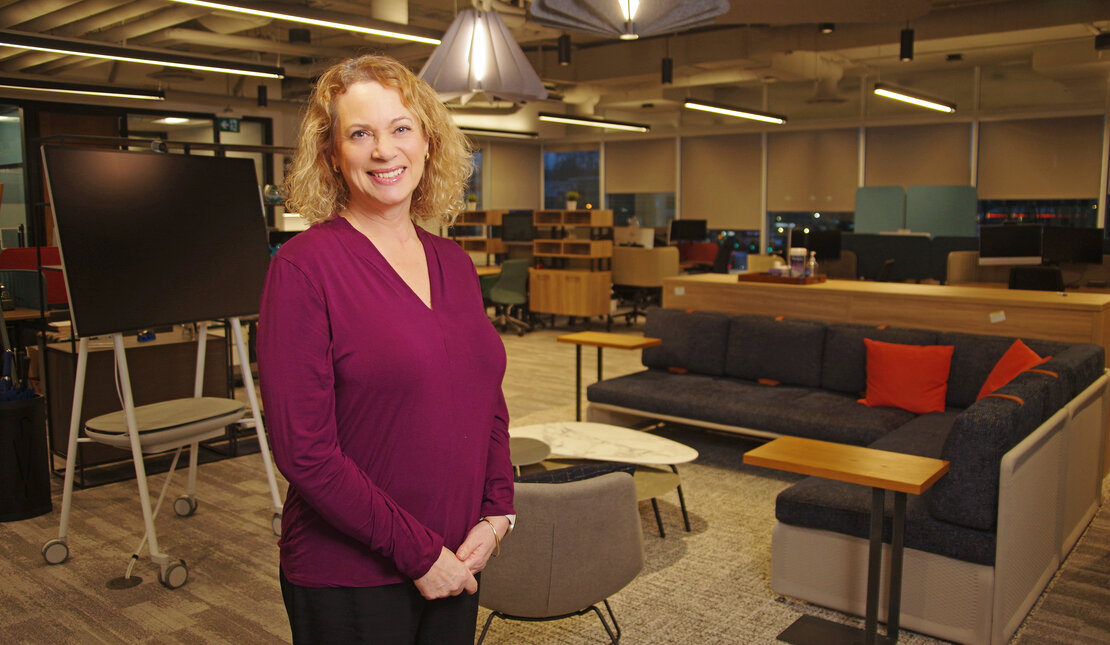Liz Bailey Connor of First West Credit Union stands for a photo, she is wearing a purple camisole and has blond curly shoulder length hair. In the background are the furniture and lighting in the building.