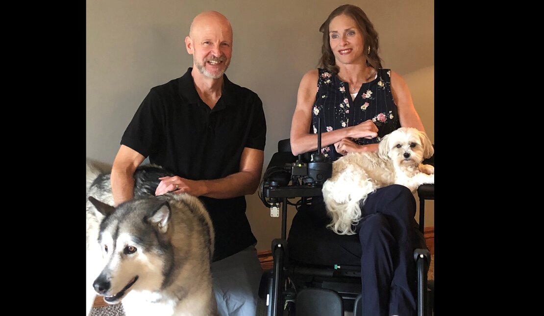 woman with shoulder length brown hari in a wheelchair, smiling with one dog on her lap and another beside her, and a man in a polo shirt kneeling next to them, smiling, with close cropped greying hair
