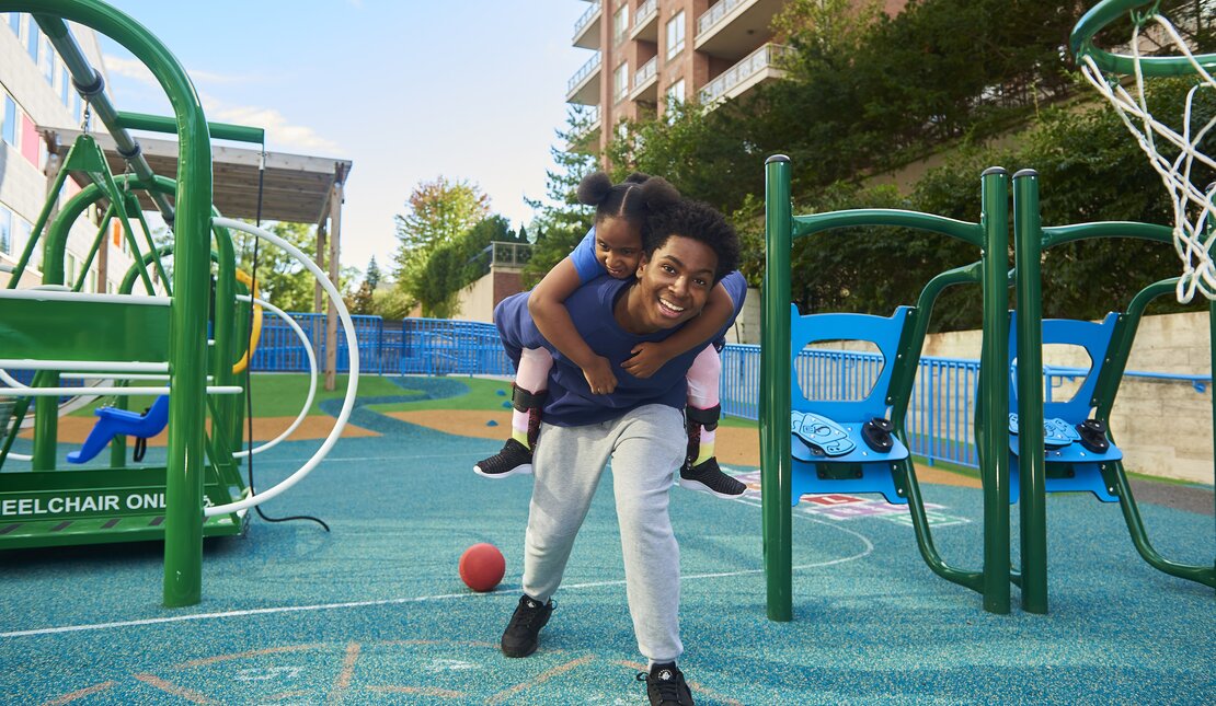 Black man and Black little girl in an accessible playground. The man has the little girl on his back and they are smiling.