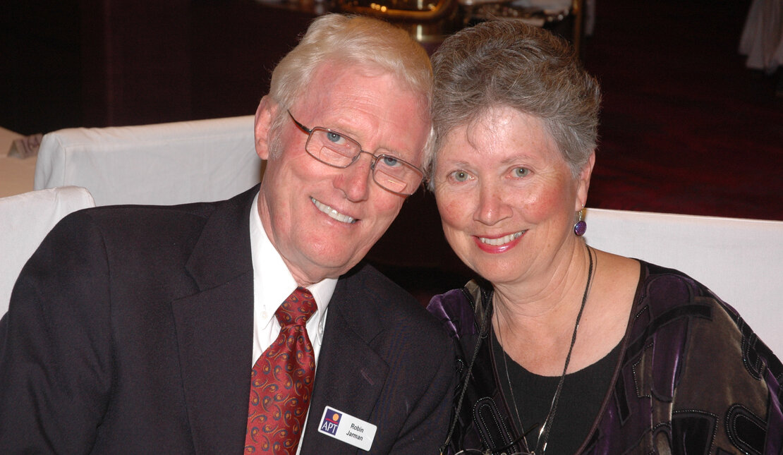 Robin and Cathryn sitting beside each other and smiling at the camera. They are older adults. Robin has white hair, glasses and is wearing a suit with a red tie. Cathryn has grey hair and is wearing purple earrings and a purple and black dress.