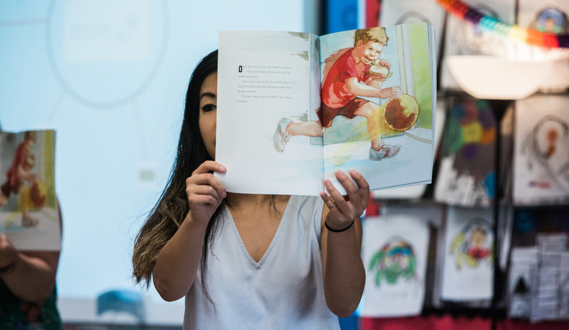 Person with long dark hair wearing a white t-shirt holding open a children's book with a colourful illustration.