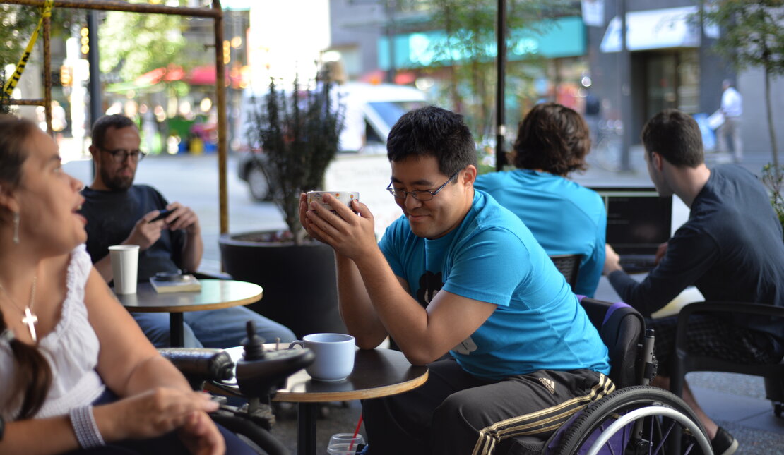 Man with black hair and glasses using a wheelchair wearing a bright blue t-shirt sitting in a cafe. He is holding a coffee cup.