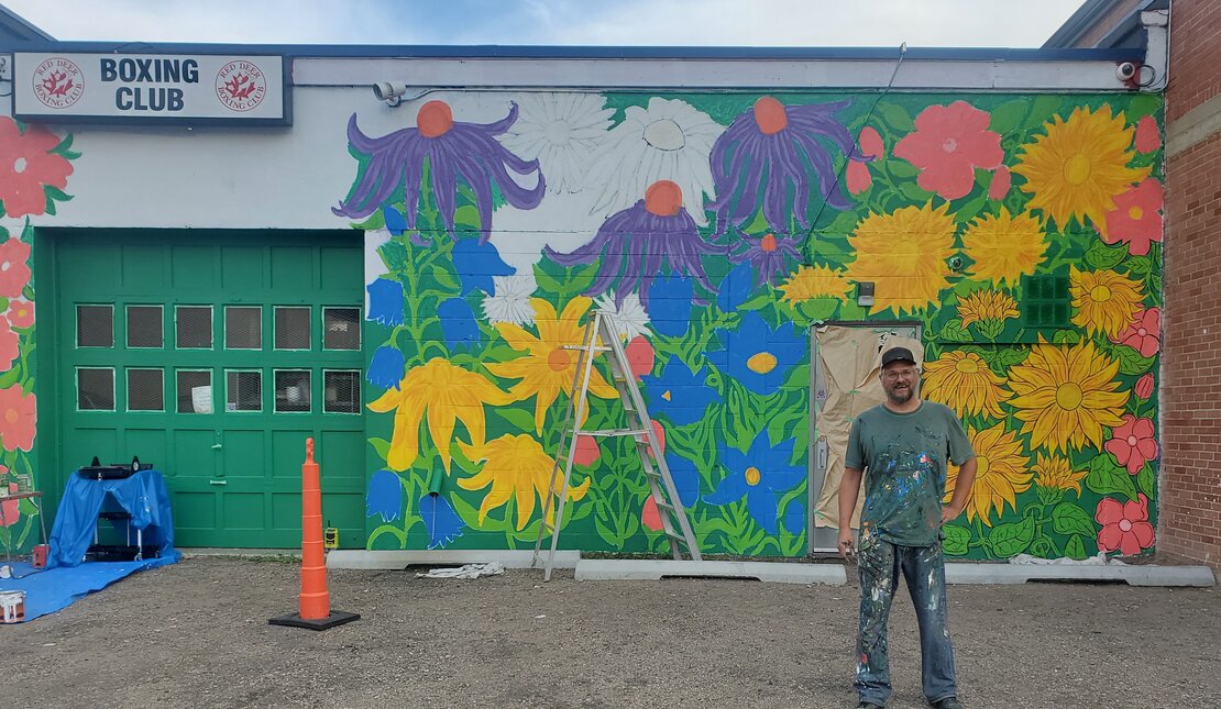 Man with clothes covered in paint standing in front of a large, colourful flower mural.