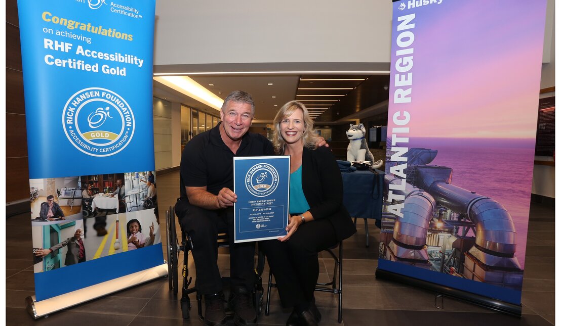 Rick Hansen (man with light brown hair) using his wheelchair holding a blue certification sign with a blonde woman who is sitting beside him. They are beside two large banners.