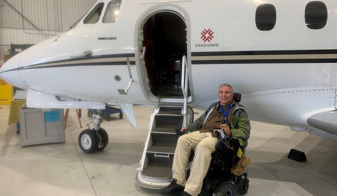 Man with light hair who is using a motorized wheelchair next to a plane. He is wearing light pants and a green and grey jacket.